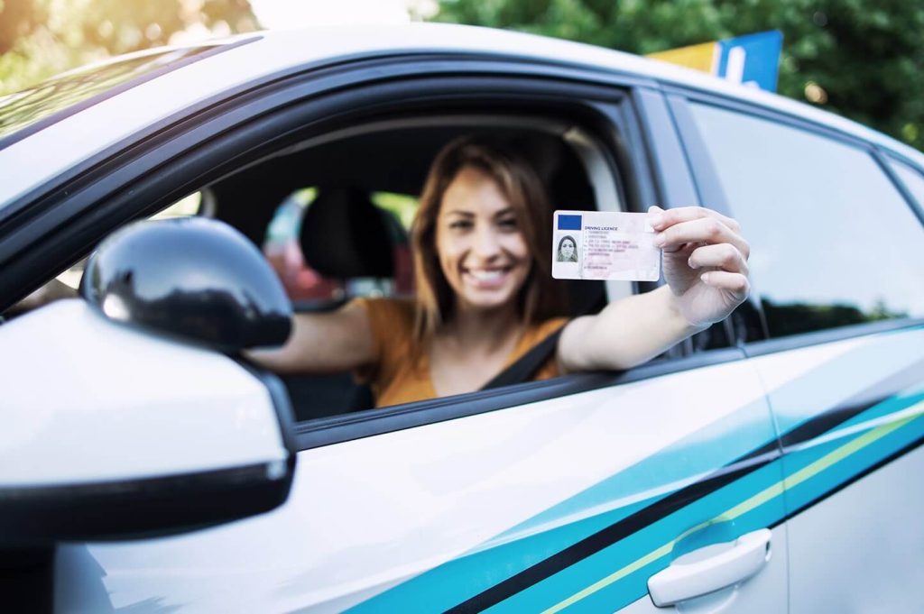 Na zdjęciu widoczna jest kobieta siedząca za kierownicą samochodu, która pokazuje prawo jazdy przez otwarte okno pojazdu. Uśmiechnięta kierowczyni prezentuje dokument bezpośrednio do kamery, co symbolizuje legalne uprawnienia do prowadzenia pojazdu oraz gotowość do jazdy